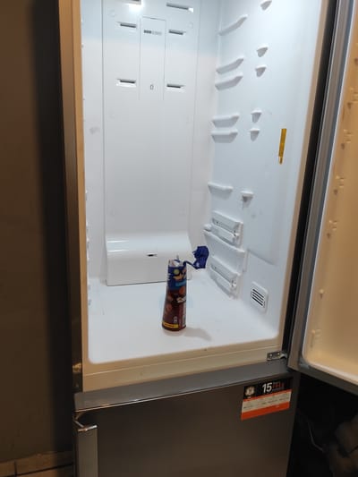 An open, empty refrigerator is pictured from the interior, with the door ajar on the right. The refrigerator's interior is white and well-lit. A single item, a tall, brown cylindrical container with a blue cap, is on the bottom shelf. The container appears to be a beverage or food product. On the lower right exterior of the refrigerator, there is an orange and white sticker with text and numbers that suggests a warranty or guarantee. The exterior of the refrigerator is silver, and the surrounding environment appears to be a dimly lit room with a dark wall to the left. No people are visible in the image. The location is Chaponost, France.
