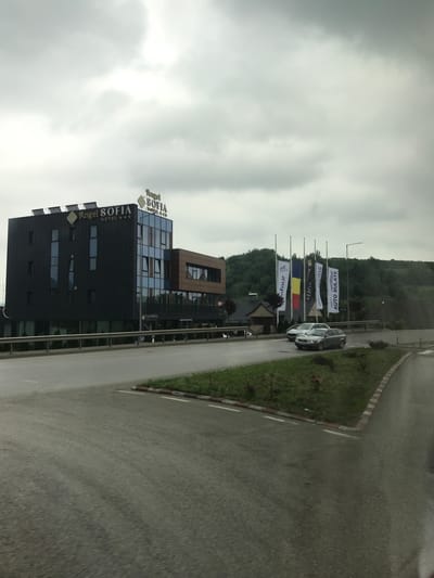 The image depicts an exterior view of the Angel Sofia Hotel, a three-star establishment, located in Fălticeni, Romania, under an overcast sky. The hotel comprises a multi-story building with a dark grey or black facade on the left, featuring multiple rectangular windows. Its flat roof is equipped with several solar thermal collectors. Adjacent to and partially integrated with this section is a modern structure with a blue-tinted glass facade, which then connects to a warmer-toned, possibly wood-paneled, section further to the right. The hotel's name, "Angel Sofia HOTEL ***", is prominently displayed in gold lettering on the top edge of the dark building, with smaller "Angel Sofia" signs visible on the glass facade and at ground level.
In the foreground, an asphalt road runs horizontally across the scene, appearing slightly damp. A grassy median strip, edged with white and red curb markings, separates different sections of the road. Two passenger cars are visible on the road in the mid-ground: a lighter-colored sedan and a darker grey station wagon, both appearing to be in motion or recently stopped.
To the right of the hotel, a line of six flagpoles stands beside the road. One clearly displays the national flag of Romania (blue, yellow, red vertical stripes). Other banners are visible, one featuring the text "romfour" and another displaying "AUTO RULATE" (Romanian for "used cars"), suggesting a nearby automotive business. A tall street light pole is positioned among the flagpoles. Behind these, a dense, dark green forested hill forms the backdrop, with smaller, residential-style buildings with dark roofs nestled at its base. The overall environment suggests a commercial area on the outskirts of a town, bordered by natural landscape. No people are visible in the scene.