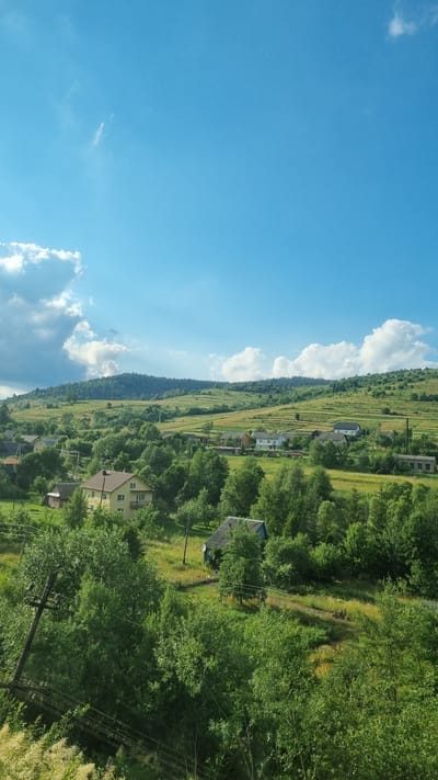 This is a vertical, eye-level, full shot of a rural landscape under a bright blue sky with scattered white clouds. The scene depicts rolling green hills dotted with houses and trees. In the foreground, lush green trees and foliage dominate, with power lines and a utility pole visible on the left. Further back, a cluster of houses can be seen nestled amongst the trees. A prominent yellow house with a balcony stands out in the middle ground. The hills in the distance are covered with vegetation and appear to be terraced in some areas, suggesting agriculture. The overall impression is of a serene, sunlit day in a village located in the city of Yavora, Ukraine.