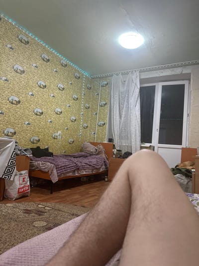 The image depicts an indoor scene, likely a bedroom, with the perspective from a person lying down. The room features yellow wallpaper with a stone pattern and what appear to be cartoonish depictions of animals in clouds. A string of blue LED lights runs along the top edge of the wall. A single, circular ceiling light fixture is illuminated.
On the left side of the room, there is a wooden-framed bed covered with a purple blanket and other bedding. To the left of the bed, a white bag with a black Greek key pattern is visible, along with a red and white shopping bag labeled "Kouffo."
In the background, to the right, is a window with white sheer curtains. The window itself appears to be a balcony door, with a dark view outside.
The foreground of the image is dominated by the legs and lower torso of a person, indicating a first-person perspective. The person's legs are light-skinned and appear to have visible hair. The background further back shows another bed with a green-patterned cover.
The overall impression is of a casual, lived-in bedroom environment. There is no specific visual information in the image to confirm its location in the city of Corlăteni, Moldova.