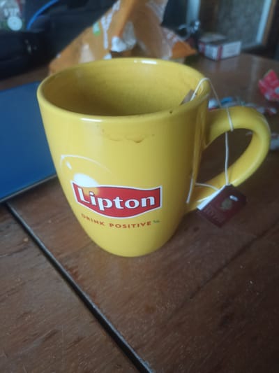 A yellow Lipton tea mug with a tea bag is placed on a wooden table. The mug has the Lipton logo and the words "DRINK POSITIVE" printed on it. In the background, there are some blurred objects that appear to be food packaging and a blue object, possibly a notebook or folder. The scene appears to be indoors. The image does not contain any information about the location in Pervomaisk, Moldova.