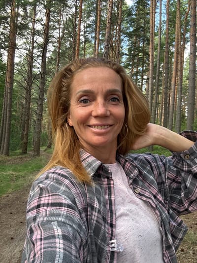 A woman with auburn hair and freckles smiles at the camera, her right arm resting behind her head. She is wearing a pink and grey plaid shirt over a pink t-shirt, and a small, clear plastic hair clip is attached to the t-shirt. The background consists of a dense forest of tall pine trees, with a dirt path visible in the foreground. The scene is set in the city of Portovoe, Russia.