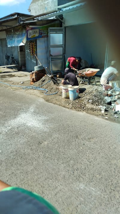The image depicts an outdoor scene likely in Cangkringsari, Indonesia, where construction work is underway. The environment appears to be a street or roadside adjacent to a building undergoing renovation or construction. In the foreground, a paved surface, possibly a road, is visible. In the mid-ground, several individuals are engaged in manual labor. One person in a red shirt and green pants is working near a wheelbarrow filled with materials. Another person in a dark shirt and cap is crouched down, seemingly mixing or handling materials in a pile of sand and gravel. A third person in a white shirt is partially visible in the background, also engaged in the work.
Various construction materials are scattered around the scene, including piles of sand and gravel, bricks, buckets, and a wheelbarrow. A blue hose is laid out on the ground, suggesting water is being used in the construction process. Behind the workers, there's a building with a corrugated metal roof and visible construction elements. A banner with text, including the word "JUARA," is displayed on the side of the building, indicating a commercial establishment. A bicycle is parked near the building. The overall impression is one of active construction work in a somewhat informal urban setting.