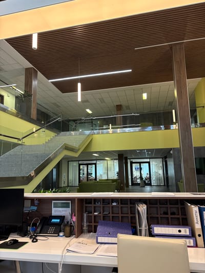 The image depicts the interior of a modern office building. The scene is dominated by a multi-level atrium with a striking wooden slatted wall and bright yellow accents. A contemporary staircase with glass railings winds through the space, connecting different floors.
On the right side of the image, a reception desk is visible. It is cluttered with office supplies, including a telephone, a computer monitor, a mouse, binders, and papers. A certificate with Cyrillic text is displayed on the wall behind the desk. There are also some decorative items and plants on the desk and shelves.
In the background, through glass doors, glimpses of other office spaces can be seen, with people working and sitting in seating areas. The overall impression is one of a clean, organized, and professional environment. The lighting suggests it is daytime. The location context is Kholmy, Russia.
Visible text includes "АНКЕТЫ" on a folder, and text on the certificate which appears to be a name and possibly an award. There is also text on some documents on the reception desk.