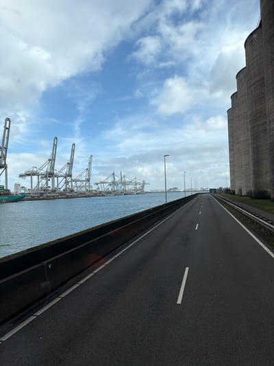 The image depicts an industrial port scene at Maasvlakte, Rotterdam, Netherlands, under a partly cloudy sky with visible blue patches. A wide expanse of water occupies the left side of the frame, bordered by a dark grey concrete barrier. Across the water, numerous large, light-colored gantry cranes are visible, indicating a container terminal where a green container ship is partially docked on the far left.

A two-lane asphalt road with white dashed lane markings extends from the foreground into the distance, occupying the center and right-center of the image. A solid white line marks the right edge of the road. Black, pole-mounted streetlights are spaced along the right side of the road. To the far right, a series of tall, dark-grey, cylindrical industrial silos with distinct horizontal banding run parallel to the road and extend into the background. A narrow strip of grass and a metal guardrail separate the road from the silos. No people or active vehicles are present in the scene.
