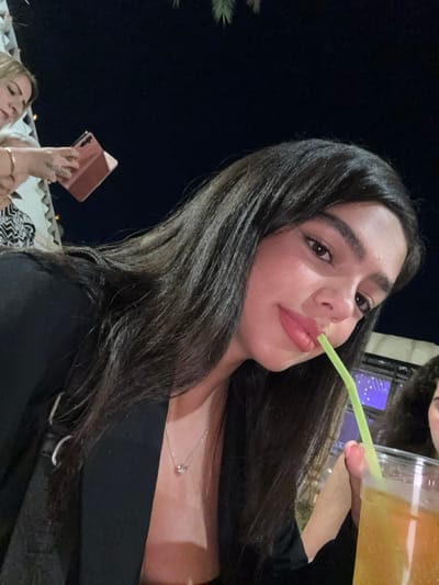 The photo shows a young woman with long dark hair, wearing a black blazer, sitting at a table outside at night, drinking from a glass of a yellowish beverage with a green straw. She is looking at the camera with a slight smile. Another young woman, wearing a black and white patterned shirt, is visible in the background, holding a pink phone. The scene is set at an outdoor restaurant or cafe in Ramla, Israel, located at 31.926 latitude and 34.873 longitude. The background is blurred, suggesting a night setting with ambient light sources.