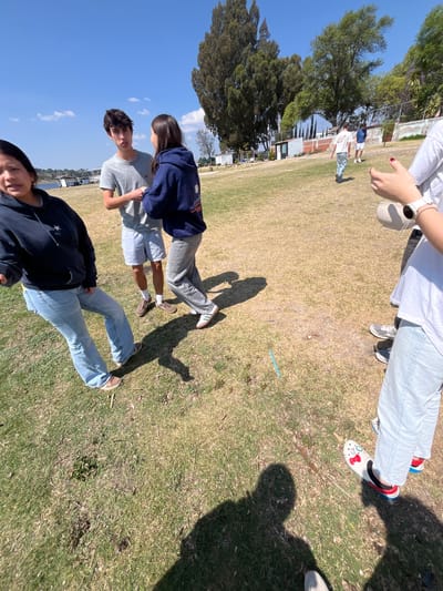 The image shows a group of people in El Oasis Valsequillo, Mexico, standing on a grassy area. One person is partially visible in the foreground, wearing a dark jacket and light blue jeans. Two people are standing next to each other in the middle ground, one wearing a grey shirt and light blue shorts and the other wearing a blue sweatshirt and light jeans. Another person is partially visible in the upper right, wearing a white shirt and light jeans. They are also holding a white cup. The environment includes trees, a blue sky, and some buildings in the background. The lighting suggests a sunny day.