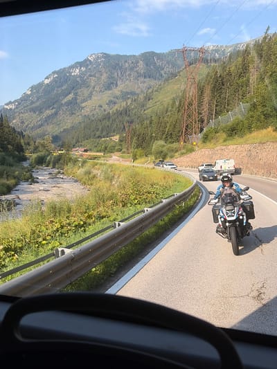 A person is riding a motorcycle on a paved road in a mountainous area. The motorcycle is white, black, and blue, and the rider is wearing a helmet and riding gear. The road curves to the right, and there are several cars and a van on the road ahead. The surrounding environment is a valley with a river, trees, and mountains. The sky is blue with some clouds. The scene appears to be captured from a vehicle moving behind the motorcycle. The location is on the SS48 road leading out of Predazzo, Italy, in the direction of the Rolle Pass.