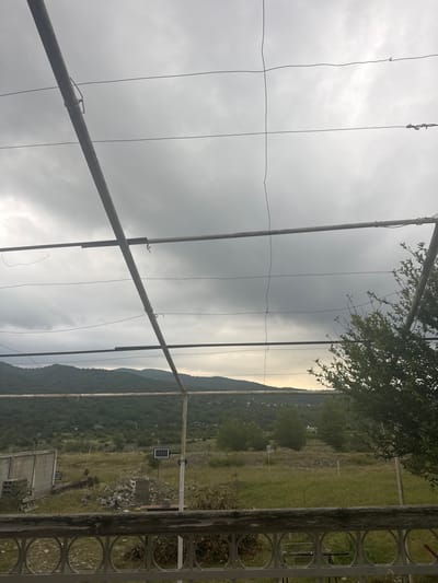 The image depicts an outdoor scene in Aragvispiri, Georgia, under a cloudy sky. In the foreground, a weathered wooden railing with circular metal accents separates the viewer from the rest of the scene. Above the railing, a metallic structure composed of horizontal and diagonal beams supports thin wires that crisscross the frame.

The middle ground features a landscape with a gently sloping hill covered in lush green trees and vegetation. A concrete wall, possibly part of a building or boundary, is visible on the left. Scattered debris, including what appears to be a pallet and rocks, lies on the ground. A small solar-powered light is mounted on a pole in the middle of a field.

The background shows a range of hills and mountains, also densely vegetated, fading into the overcast sky. The overall impression is one of a rural or semi-rural setting. No people are present in the image.