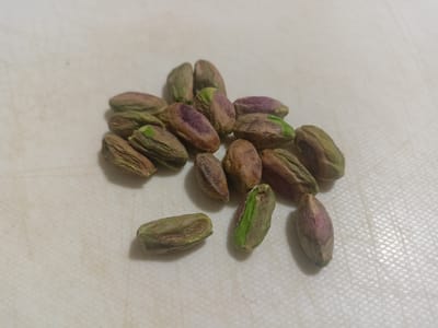 The image shows a pile of unshelled pistachios on a textured white surface. The pistachios vary in color, with some exhibiting shades of green, brown, and purple. The lighting suggests a close-up shot, possibly taken indoors. There is no discernible urban environment or indication of the city of Grudziądz, Poland. The scene is static, with no action or interaction depicted.
