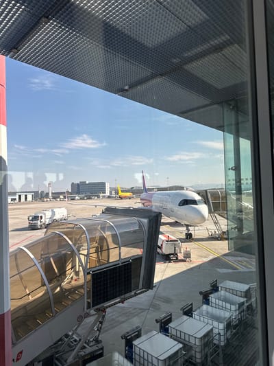 The image depicts an airport tarmac scene viewed through a large window, likely from a terminal building, located at EuroAirport Basel Mulhouse Freiburg, which serves Saint-Louis, France. The sky is bright blue with scattered white clouds.

In the immediate foreground, a textured, grid-patterned ceiling panel is visible across the top of the frame. A thick, red and white striped vertical column is present on the left. Faint reflections of interior elements, including indistinct human silhouettes, are visible on the glass. In the lower right, a stack of white intermediate bulk containers (IBCs) is present.

The midground features an active tarmac. A white passenger jet, identifiable by its purple tail and partial fuselage livery (likely Wizz Air, but text is indistinct), is parked, with a mobile jet bridge connected to its forward door. This jet bridge is enclosed with brown-tinted transparent panels and has black solar panels mounted on its side. A white ground service vehicle is positioned near the nose of the parked aircraft. To the left of the jet bridge, a white tanker truck with a visible driver is in motion on the tarmac. Further back on the tarmac, a yellow aircraft, potentially a cargo plane with DHL markings, is also parked.

The background shows multiple airport structures, including multi-story buildings and what appears to be an air traffic control tower, under bright daylight conditions, casting shadows on the paved tarmac.