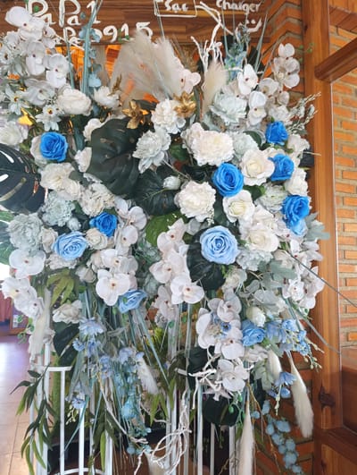 This is a full-shot, eye-level, vertical photograph of a large floral arrangement positioned in front of a wooden structure with a brick wall in the background. The floral arrangement is predominantly composed of white and pale blue flowers, with some dark green foliage and light-colored Pampas grass plumes. A wooden sign with white lettering is partially visible at the top of the arrangement, displaying text that appears to include "Pa 1 R 915" and "sa Charger". The arrangement is set against a backdrop that suggests an indoor or covered outdoor setting, possibly a wedding venue or a restaurant, given the wooden beams and brickwork, and the floor tiles visible at the bottom of the image. The scene is well-lit, with natural light illuminating the flowers and surrounding area. No people are present in the photograph. The overall impression is one of elaborate decoration, likely for an event or as a permanent fixture. The location is identified as Lương Sơn, Vietnam.