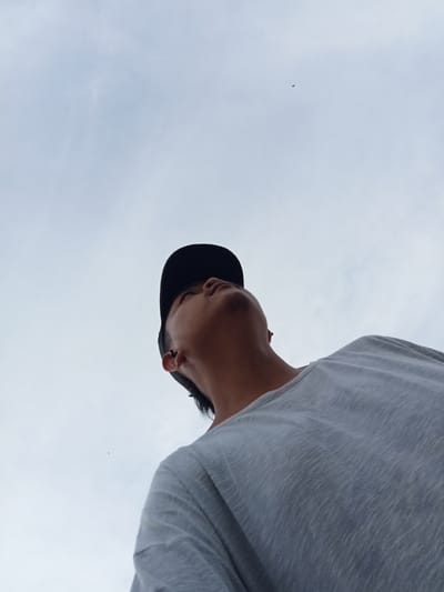 A low-angle shot captures a young person from the shoulders up, wearing a light grey t-shirt and a black baseball cap. They are looking upwards, their face angled towards the sky. The background is an overcast sky with subtle cloud formations and two small dark specks that appear to be birds in flight. The image is taken outdoors, possibly in Kualu, Indonesia, given the context.