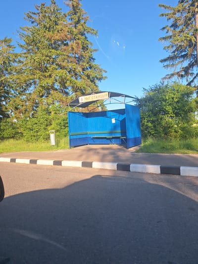 The image shows a bus stop shelter on a sunny day. The shelter is blue with a metal roof and has a bench inside. There are trees and greenery behind the shelter. A trash can is located to the left of the shelter. The bus stop is situated next to a paved road with a curb marked with black and white stripes. A sign above the shelter reads "А ЧЫГ. СТАНЦЫЯ", which translates to "Bus Stop" in Belarusian. The location is identified as Цімкавіцкі сельскі Савет, Belarus.