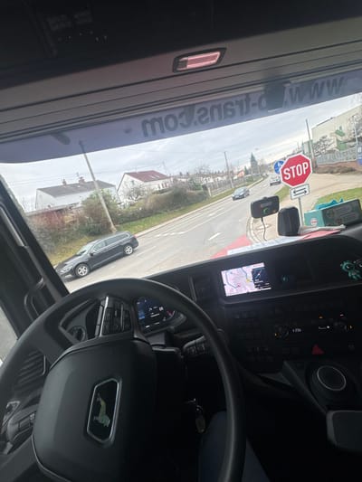 The image provides a first-person view from the driver's position inside the cabin of a stationary heavy goods vehicle, likely a DAF truck, at an intersection in Friedberg, Germany. The steering wheel, featuring a prominent emblem, occupies the lower left, with the dashboard displaying a digital speedometer indicating 0 km/h and an integrated navigation screen showing a local map. Vehicle keys hang from the ignition or a hook. On the dashboard, a black dash-mounted device with a green indicator light is visible, along with a red box (possibly a first-aid kit) and a light blue item on the passenger side. A clear reflection of 'www.d-trans.com' is visible inverted on the upper windshield. Outside the vehicle, a two-lane asphalt road extends forward. A red octagonal 'STOP' sign with a white border and an associated priority sign stands on the right side of the road. A black station wagon is positioned on the left side of the road, facing away from the viewer. Several residential houses with sloped roofs are visible on both sides of the road, interspersed with trees and bushes. The sky is overcast, and other vehicles can be observed in the distance on the road. No people are explicitly visible.