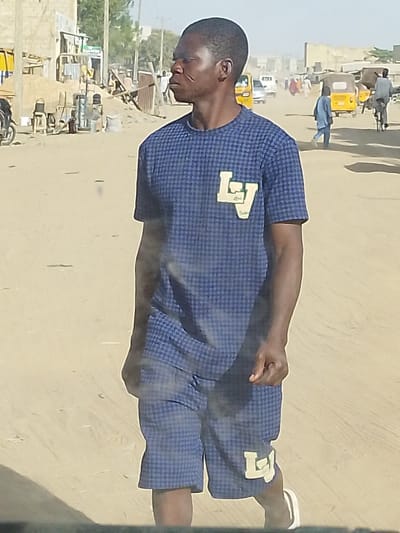 The image captures a full-length shot of a dark-skinned man walking on an unpaved, dusty road under clear daylight. He is positioned slightly off-center to the left, facing towards the left side of the frame, with his head turned slightly upward.
The man has short, dark hair and prominent scarification marks on his left cheek and temple area. He is wearing a matching two-piece outfit consisting of a short-sleeved t-shirt and shorts, both in a dark blue color with a small, light-colored checkered or houndstooth pattern. On the left chest area of his shirt, there is a prominent light-colored patch or appliqué, stylized to resemble the letters "LV". A similar "LV" patch is visible on the left leg of his shorts. His right arm is bent, and his hand is visible, while his left arm is mostly obscured. He is wearing light-colored shoes with dark soles, possibly sneakers or sandals.
The background depicts a bustling, dusty street scene. On the immediate left, there are buildings made of light-colored block or concrete, with some construction materials like piles of sand or dirt, and wooden planks. A motorcycle is partially visible on the far left. The road itself is wide and unpaved, appearing to be dirt or sand. Further down the road, several yellow auto-rickshaws (tuk-tuks) are scattered, along with other cars in the distance. Multiple people are visible in the mid-ground and background, including one person in blue walking towards the right, and another person on a bicycle on the far right. The sky is bright and clear, indicating a sunny day. The overall lighting suggests strong sunlight, casting some subtle shadows. There are some smudges or dust particles visible across the image, potentially on the camera lens or a window through which the photo was taken, giving a slightly hazy effect over the man's lower body.