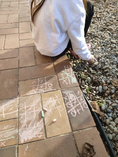 An outdoor scene in Aubagne, France, depicts a person, observed from a rear-side perspective, in a kneeling or squatting posture. The individual wears a white long-sleeved top and dark trousers, with brown hair visible over the right shoulder and a glittery pink shoe on the right foot. Their right hand is positioned near a gravel bed to the right of the paved area. The ground consists of reddish-brown rectangular paving stones, arranged in a staggered pattern, adjacent to the gravel bed. Multiple Tic-Tac-Toe grids, drawn with white, light pink, and light green chalk, are visible on the pavers. Three broken pieces of chalk—one green and two pink—are scattered on the paved surface. The gravel bed comprises small, smooth, multi-colored pebbles, interspersed with sparse green vegetation and dried brown leaves. A thick black hose or cable runs along the boundary between the paving and the gravel. The scene is illuminated by direct sunlight, casting shadows.