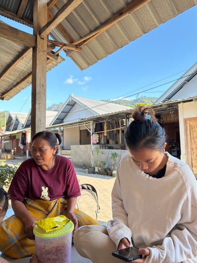 In Desa Pemenang Barat, Indonesia, a woman in a maroon t-shirt and a yellow plaid sarong sits on the ground, looking to her left. In front of her is a clear plastic container filled with chopped red and white items, topped with a yellow packet. Beside her, a young woman in a cream-colored sweatshirt and light-colored pants is looking down at a black smartphone she holds in her hands. Behind them, there are simple structures with corrugated metal roofs, laundry hanging on racks, and potted plants. Mountains are visible in the distance under a clear blue sky.