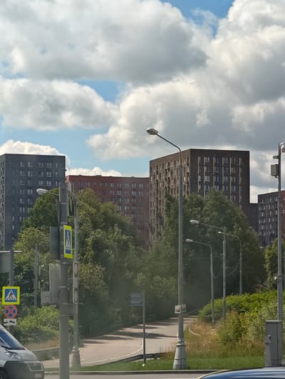 The image depicts a street scene in Нефтемаш Снт, Russia, on a cloudy day. In the background, there are several multi-story apartment buildings of varying architectural styles and colors, including gray, reddish-brown, and dark brown brick. Lush green trees and dense foliage line the street and surround the buildings. A paved road curves through the scene, bordered by grass and other vegetation. Several streetlights are visible, along with traffic signals and road signs. One prominent sign is a pedestrian crossing sign. A portion of a silver vehicle is visible in the lower left corner of the image. The sky is filled with large, white, fluffy clouds interspersed with patches of blue sky.