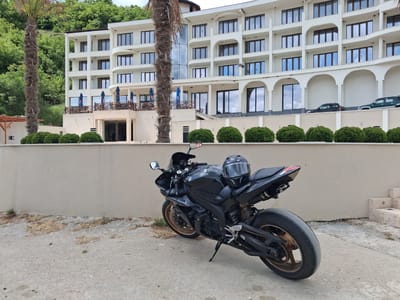 A black Yamaha motorcycle with a dark helmet resting on the seat is parked on a concrete surface in front of a light-colored wall. Behind the wall is a neatly trimmed row of green bushes, followed by a multi-story white building with many windows. The building has a section with arched openings at ground level. Two vehicles, a black car and a green SUV, are visible parked behind the bushes. Two palm trees are positioned in front of the building, with a green hillside and forest visible in the background. The scene appears to be located in an outdoor area near the city of Pisanets, Bulgaria.