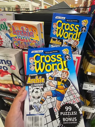 This photograph shows a selection of magazines or puzzle books displayed on a store shelf. Dominating the view are copies of "Cross Word" puzzle books featuring Archie and his friends. The books are blue with the title prominently displayed in yellow lettering. One book features an illustration of Archie kicking a soccer ball. Additional titles visible include "Archie Comics Digest Summer Fun", and another featuring desserts on the cover. A hand is visible near one of the "Cross Word" puzzle books, appearing to be holding it. Several price tags are attached to items on the shelf above the books. The location is in a store in Key Largo, United States of America.