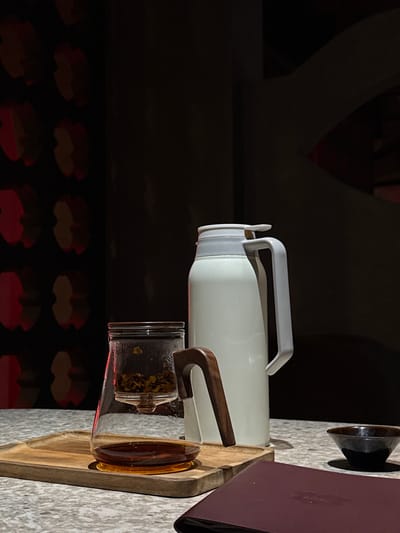 A tea set is arranged on a textured surface, possibly a table in a cafe or restaurant. The setup includes a clear glass teapot with dried tea leaves in the upper infuser and brewed tea in the bottom chamber. Next to it, a tall, white thermos with a grey lid and handle stands prominently. To the right, a small, dark ceramic bowl sits on the surface. The background features a dark wall with a repeating pattern of red and light brown shapes, creating an atmospheric and dimly lit environment. The lighting casts shadows that add depth to the scene. The specific location within the city of Бородино, Russia, is not discernible from the visual information provided. There are no people or animals present in the image, and no actions or interactions are depicted.