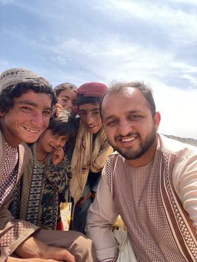 The image depicts a group of individuals in an outdoor setting, likely Ashoqah, Afghanistan, based on cultural attire and landscape features. There are five individuals visible. The man on the right, dressed in a light-colored traditional Afghan shirt, is taking the photo. Next to him are three younger individuals, one wearing a maroon cap and another with a tan scarf. The girl has her fingers in her mouth, with a floral shirt and beaded necklace. On the left is a man wearing a patterned Afghan cap and traditional clothing smiling. The background features a clear sky and what appears to be a barren, mountainous terrain with hazy outlines of mountain peaks in the distance.