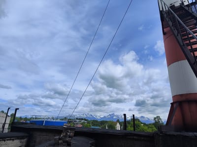 The image shows a view of Milkovo, Russia, with a lighthouse-like structure in the foreground on the right. The structure is red and white with a black ladder. In the mid-ground, there are several buildings, including a blue-roofed structure and some houses, surrounded by trees. In the background, snow-capped mountains are visible under a cloudy sky. The foreground consists of a dark, possibly tar-covered roof with some brickwork. Power lines stretch across the sky. There are no people visible in the image.