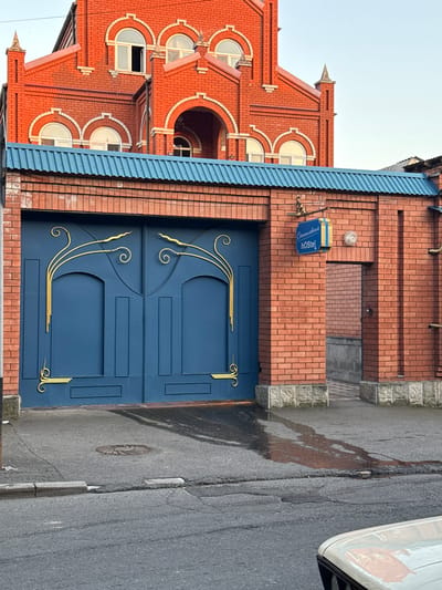 This image shows a large, dark blue metal gate with ornate gold swirling designs, set within a red brick wall. The gate is imposing and appears to be the entrance to a property. Above the gate, the roof of an adjacent structure is visible, made of corrugated blue metal. The brick wall also has a smaller, open doorway to the right of the gate, leading to an interior courtyard or alleyway. A blue sign with yellow lettering is affixed to the brick wall above this doorway, which reads "Славянскaя hOstel".
The architecture of the building visible behind the gate and wall is distinctive, featuring red brick construction with decorative white accents around arched windows and a prominent central archway. The building has a steeply pitched roof with dormer windows. The overall style suggests a potentially older or historically inspired design.
The foreground of the image is a paved road, appearing somewhat worn with cracks and a circular manhole cover visible. There is a patch of wetness on the pavement near the gate, possibly from recent rain or a leak. A portion of a light-colored vehicle is visible in the bottom right corner of the frame. The sky is a pale blue, indicating it is likely daytime. The scene appears to be an exterior urban or semi-urban environment. The location is identified as Sulak, Russia.