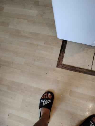 The image features a person wearing black Adidas slides on a light wooden floor. The person has two slides on. A corner of a white object is visible on the top left side of the image. The wooden floor is composed of parallel, rectangular planks and has a dark wooden frame around it.