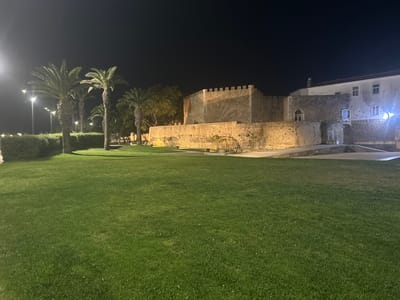 In Lagos, Portugal, a nighttime scene features a meticulously manicured green lawn in the foreground. Behind the lawn, a stone wall with crenellations marks the perimeter of a historical structure, possibly a fortress or castle, illuminated by warm, ambient lighting. Palm trees stand tall to the left of the frame, their fronds silhouetted against the dark night sky. Streetlights are visible in the distance, casting a soft glow. The architecture suggests a blend of ancient and more modern elements, with a white building visible to the right of the stone structure. The overall impression is one of historical significance and peaceful evening ambiance.