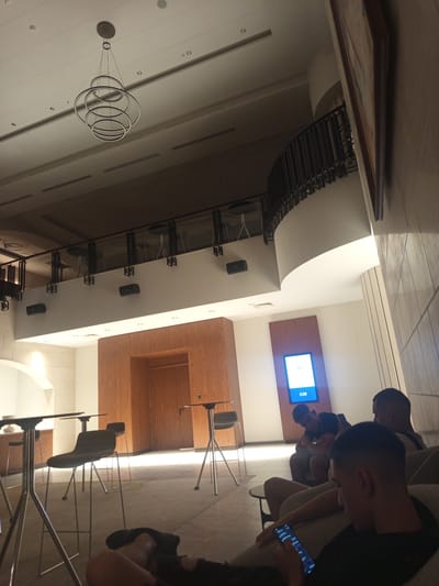 The image depicts an indoor scene, possibly a lounge or waiting area, with a modern aesthetic. In the foreground, a person is partially visible, seated and looking at their mobile phone. To the left, several high-top tables and stools are arranged, with one stool prominently featured. In the background, a large wooden-paneled wall with an inset digital screen displaying the time "0:28" is visible. Three other individuals are seated on a couch, also engaged with their phones. The ceiling features a contemporary chandelier and recessed lighting. A balcony with a railing is visible on the upper level. The overall impression is one of a contemporary public space designed for relaxation or waiting. There are no discernible elements in the image that would allow for a specific determination of its location within Razliv, Bulgaria.