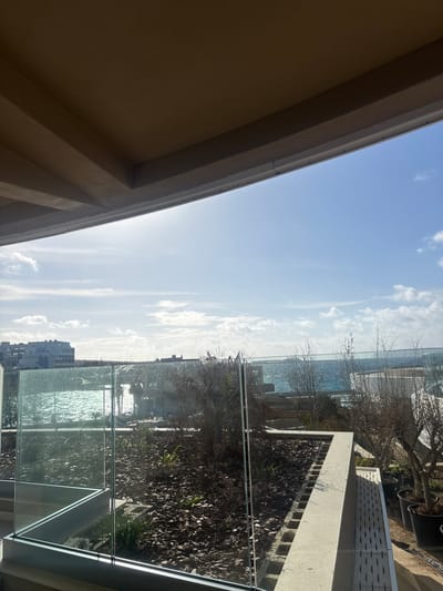 View from a balcony with a glass railing overlooking the Mediterranean Sea. In the background, buildings are visible along the coast, suggesting an urban area. The balcony has a planter box filled with mulch and a few plants. Part of a building's exterior, possibly the balcony's ceiling, is visible in the foreground, obscuring the upper left corner of the view. The sky is blue with scattered clouds, indicating clear weather. Location is suggested to be Mellieħa, Malta.
