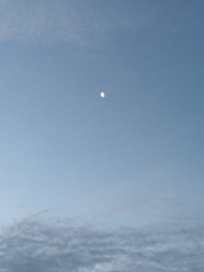 The image shows a bright, crescent-shaped object in a clear, pale blue sky, positioned slightly above the center of the frame. The object appears to be illuminated from the left, with a distinct curved shadow. Below this object, the sky transitions to a hazy, muted blue, with blurred, cloud-like formations at the bottom of the image. The overall scene is overcast and somewhat muted in color. There are no people or man-made objects visible in the image. The location of the photograph is the city of Soborne, Ukraine.