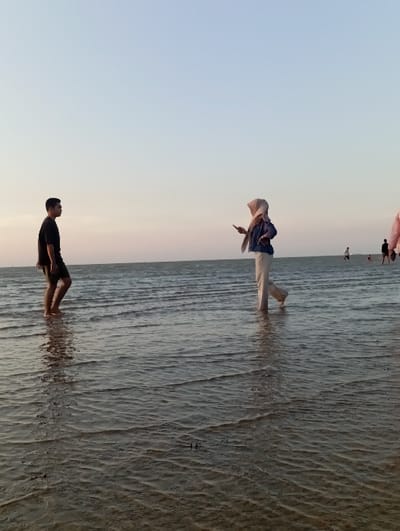 The image displays a shallow coastal area in Tritunggal, Indonesia, under a twilight sky that transitions from light blue overhead to a pale pink-orange at the horizon. The foreground is comprised of rippled, shallow water with a visible dark, sandy-muddy seabed. Two individuals are positioned in the midground, standing barefoot in the water. On the left, a male is seen in right profile, wearing a black short-sleeved shirt and dark shorts, with water reaching his shins. On the right, a female is depicted from her left side, attired in a light-colored hijab, a blue long-sleeved top, and light-colored long pants, with water at her ankles. She holds a dark-colored smartphone in her slightly raised left hand. The two individuals are separated by several feet. In the background, multiple additional human figures are discernible standing in the water, appearing as dark silhouettes against the distant horizon.