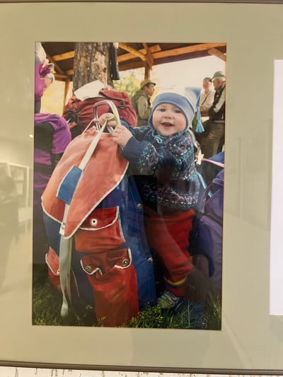 An image displays a smiling baby, appearing to be an infant or young toddler, situated outdoors. The baby is positioned centrally, looking directly at the camera with a wide, joyful smile, revealing a few teeth. They are wearing a light blue knitted beanie with a tassel, a dark blue sweater with a colorful pattern (possibly fair isle style), and red pants.
The baby's hands are holding onto a light red fabric item that resembles a climbing rope bag or a gear sling, with a white strap and a blue patch, and metal carabiners visible near its top edge. This item rests on a larger blue fabric surface, possibly a mat or another piece of outdoor gear. Behind the baby, green grass is visible to the right.
In the blurred background, several adults are partially visible, one wearing a hat and another with a backpack. A wooden structure, possibly part of a building or shelter, is also present, suggesting a rustic or natural setting. Other outdoor gear, including red and purple backpacks, can be seen in the background. The overall scene conveys a cheerful outdoor activity, likely in a natural or resort-like environment such as Sanatoriya Yumatovo Imeni 15-Letiya Bassr, Russia, during daytime under soft, natural light. No discernible text is visible within the image.