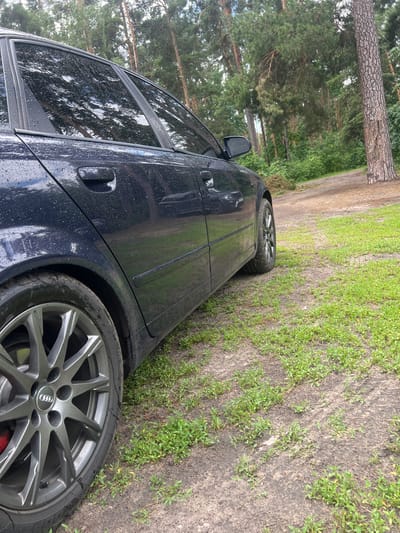 An Audi A4 Avant, dark blue, with water droplets on its surface, is parked on a dirt and grass path in a wooded area. The car is viewed from a low angle, focusing on the rear wheel and the side of the vehicle. The background consists of tall pine trees and greenery. The scene is likely outdoors in Lyutizh, Ukraine.