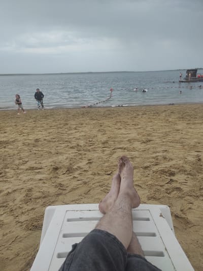 The image shows a first-person perspective of a person relaxing on a beach. The person's legs, with dark grey shorts and bare feet, are propped up on a white plastic lounge chair. The chair is situated on a sandy beach, with the sand showing footprints and natural textures. In the background, the sea stretches out under a cloudy sky. Along the shoreline, several people are visible. A young girl in a light-colored top and dark shorts is standing in the shallow water to the left, appearing to hold something yellow. A person in dark clothing stands behind her. Further out in the water, a group of people are swimming or wading, with some wearing life jackets. A small floating platform or dock with people standing on it is visible on the right side of the water. Buoy markers with red and white stripes are present in the water, indicating a designated swimming area. The environment suggests a public beach on a lake or sea, likely in Kotyubok, Kazakhstan, given the context. The overall atmosphere is one of leisure and relaxation.