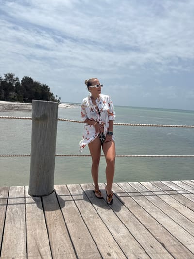 A female subject stands on a weathered wooden pier in Michamvi Kae, Tanzania. She is positioned slightly left of center, facing right with her head turned further right, looking out over the calm, greenish-blue ocean water. Her light-colored hair is styled in an updo, and she wears white rectangular sunglasses, large earrings, a thick gold-colored chain necklace, a watch on her left wrist, and a bracelet on her right wrist. She is dressed in a loose, white, long-sleeved button-up cover-up adorned with red and blue starfish and marine life patterns, open at the front to reveal a dark, leopard-print bikini bottom. Her footwear consists of multi-colored flip-flops. To her immediate left, a thick, round wooden post is integrated into the pier, supporting two parallel light-colored ropes that form a railing extending horizontally over the water. In the distant left background, a sandy shoreline is visible, backed by dense tropical vegetation and several structures. The sky overhead is light blue with scattered white clouds, and direct sunlight casts shadows on the pier's wooden planks.