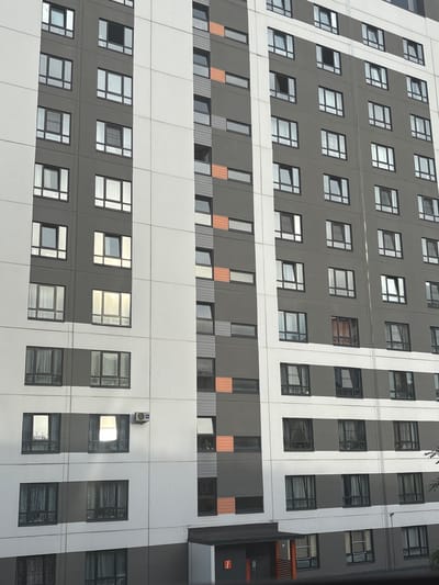 The image shows a modern apartment building in Kamenka, Russia. The building has a gray and white facade with numerous windows. Some windows are open, and some are closed. An air conditioning unit is mounted on the side of the building. The sky is overcast. The image appears to have been taken from a lower angle, looking up at the building.