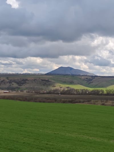 The image shows a landscape with a large green field in the foreground. Behind the field, there's a line of vegetation and a low-lying area. In the mid-ground, there are rolling hills with patches of green and bare trees, with a few buildings visible. In the background, a prominent, conical mountain dominates the scene, set against a cloudy sky. The scene appears to be in the city of Юца, Russia.