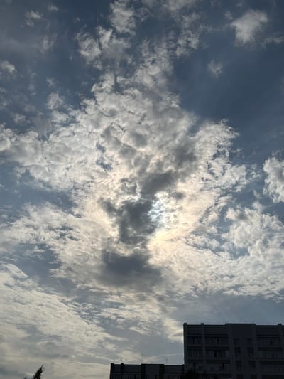 The image captures a cloudy sky above buildings in Noyabrsk, Russia. The clouds are varied, with some areas appearing bright and illuminated by the sun peeking through, while other sections are darker and more dense. In the lower portion of the image, the tops of two multi-story buildings are visible. The building on the right is larger and appears to be a modern structure with numerous windows. The building on the left is partially visible and seems to be of a similar style. There are also faint overhead wires stretching across the sky in the lower left quadrant. The overall impression is a typical urban sky scene with atmospheric conditions.