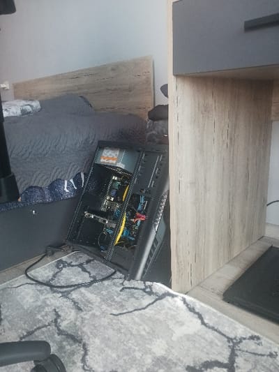The scene depicts an interior room in Velichkovo, Bulgaria. In the center, a black computer tower, with its left side panel removed, rests on a light grey rug featuring dark grey irregular patterns. The internal components, including circuit boards, a visible fan, and electrical wiring in yellow, blue, and red, are exposed. A white warning label is affixed inside the case. To the left, a bed is visible with a grey quilted comforter and a blue and white patterned sheet, positioned beneath a light wood-grain headboard against a light grey wall. To the right, a light wood-grain desk or shelf unit, featuring a dark grey drawer front, partially obscures the computer tower. A black cable lies across the rug in the foreground. A black rectangular object, possibly a laptop or device stand, is situated on the floor beneath the desk unit. No individuals are present.