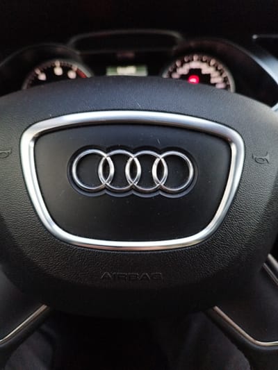 The image shows a close-up of a black leather steering wheel with the Audi rings logo in the center. The steering wheel also has silver trim around the logo and on the spokes. The word "AIRBAG" is embossed on the lower part of the steering wheel. In the background, blurred images of car dashboard instruments are visible, including a speedometer with a red light illuminated. The lighting suggests this is an indoor shot, likely within a car. There are no people visible in the image. The scene is a generic interior of a car. There is no information to suggest it is located in Schiffdorf, Germany.