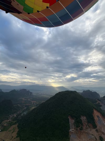 An aerial, wide-angle shot captures a breathtaking sunrise over a lush, mountainous landscape in Hong Ngern Village, Laos. The foreground is dominated by the vibrant, multicolored fabric of a hot air balloon, partially obscuring the sky. The sky itself is a canvas of soft, diffused light, with rays of sun piercing through scattered clouds. Below, a panorama of rolling green hills and dramatic, karst formations unfolds. In the distance, a hazy valley reveals a small village nestled among the trees, with patches of cultivated fields. Two other hot air balloons are visible in the distance, adding to the sense of adventure and tranquility. The overall scene evokes a feeling of awe and wonder, as the natural beauty of Laos is illuminated by the dawn.