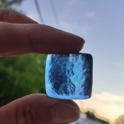 A close-up shot captures a hand holding a square, iridescent blue stone. The stone exhibits swirling patterns and reflects light, creating a mesmerizing effect. The background is blurred, showing hints of green foliage and a soft blue sky with some white clouds, suggesting an outdoor setting during daylight. The focus is clearly on the stone and the fingers holding it. The person's fingers appear to be an adult's, with visible skin texture. The overall impression is one of gentle holding and appreciation of the object. There is no visible text in the image. The hints provided suggest the scene is outdoors with a focus on the sky and foliage, and also highlight the texture of the hand and the detail of the stone.