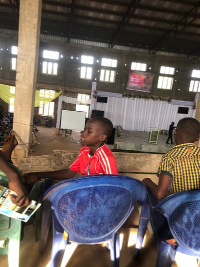 The image was taken indoors at an event in the city of Nteje, Nigeria. Two young men are visible near the camera, seated in blue plastic chairs. The person closer to the camera is wearing a red shirt with white stripes. Behind them, a stage area is decorated with white fabric and flowers, and a banner hangs between windows above the stage. A screen is visible on the left side of the stage area. People are seated in rows of chairs facing the stage. A partially obscured person in the foreground holds a brochure. The building has high ceilings with exposed beams and multiple windows, allowing sunlight to illuminate the interior.