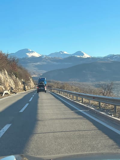 The image depicts a two-lane asphalt road in Miločani, Montenegro, observed from the perspective of a vehicle moving forward. The roadway features solid white lines marking the shoulders and dashed white lines separating the lanes. Ahead in the right lane, a dark-colored sport utility vehicle (SUV) or minivan is present, followed by a large blue commercial truck, possibly a dump truck or flatbed with a high-sided cargo area. To the left of the road, a rocky embankment rises, characterized by sparse, dry vegetation and exposed rock faces. To the right, a silver metal guardrail runs along the shoulder, beyond which is a downward sloping terrain with sparse, dry vegetation and leafless trees. In the background, multiple mountain ranges are visible, with the highest peaks covered in snow. The sky above is clear and bright blue. Long shadows extend diagonally across the right lane, indicating a low sun angle.
