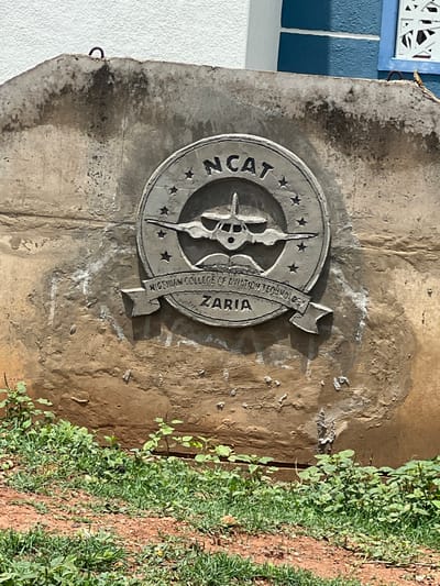 The image shows a weathered concrete wall with a circular emblem mounted on it. The emblem features an airplane in the center, surrounded by stars and text that reads "NCAT NIGERIAN COLLEGE OF AVIATION TECHNOLOGY ZARIA". The wall itself is textured and stained, with some white markings and discolored patches. In front of the wall, there is a patch of green grass and some small plants growing from the reddish soil. In the background, a white building is visible with blue accents and a white decorative window grill. The scene appears to be outdoors, under natural light. The location is identified as Basawa, Nigeria.