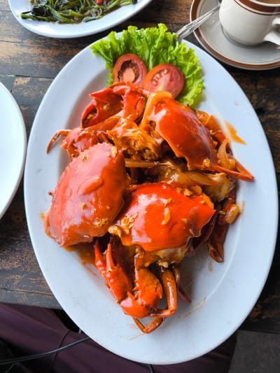 Captured in Jembrana, Indonesia, the image presents a detailed view of a dining experience, centered on a substantial meal served on a rustic wooden table. The primary subject is a large white oval platter prominently positioned in the middle, laden with multiple bright orange-red cooked crabs. These crabs are generously coated in a glossy, thick reddish-orange sauce, which contains visible lighter-colored flecks, likely chopped garlic or onion. The dish is garnished on its upper-left side with two circular slices of red tomato and several vibrant green lettuce leaves.
In the upper-left background, a segment of another white plate is visible, holding a portion of green leafy vegetables, possibly stir-fried kangkung (water spinach), submerged in a dark, reflective sauce. Towards the upper-right, a cream-colored ceramic teacup with brown rim detailing rests on a matching saucer, accompanied by a silver spoon. The various dishes are placed on a wooden table characterized by distinct grain patterns and variations in dark brown tones, suggesting a natural and possibly weathered surface. In the lower-left corner of the frame, a segment of a human's lap or leg, clad in dark maroon or purple fabric, is partially visible, with a black electrical cable or wire extending across it.